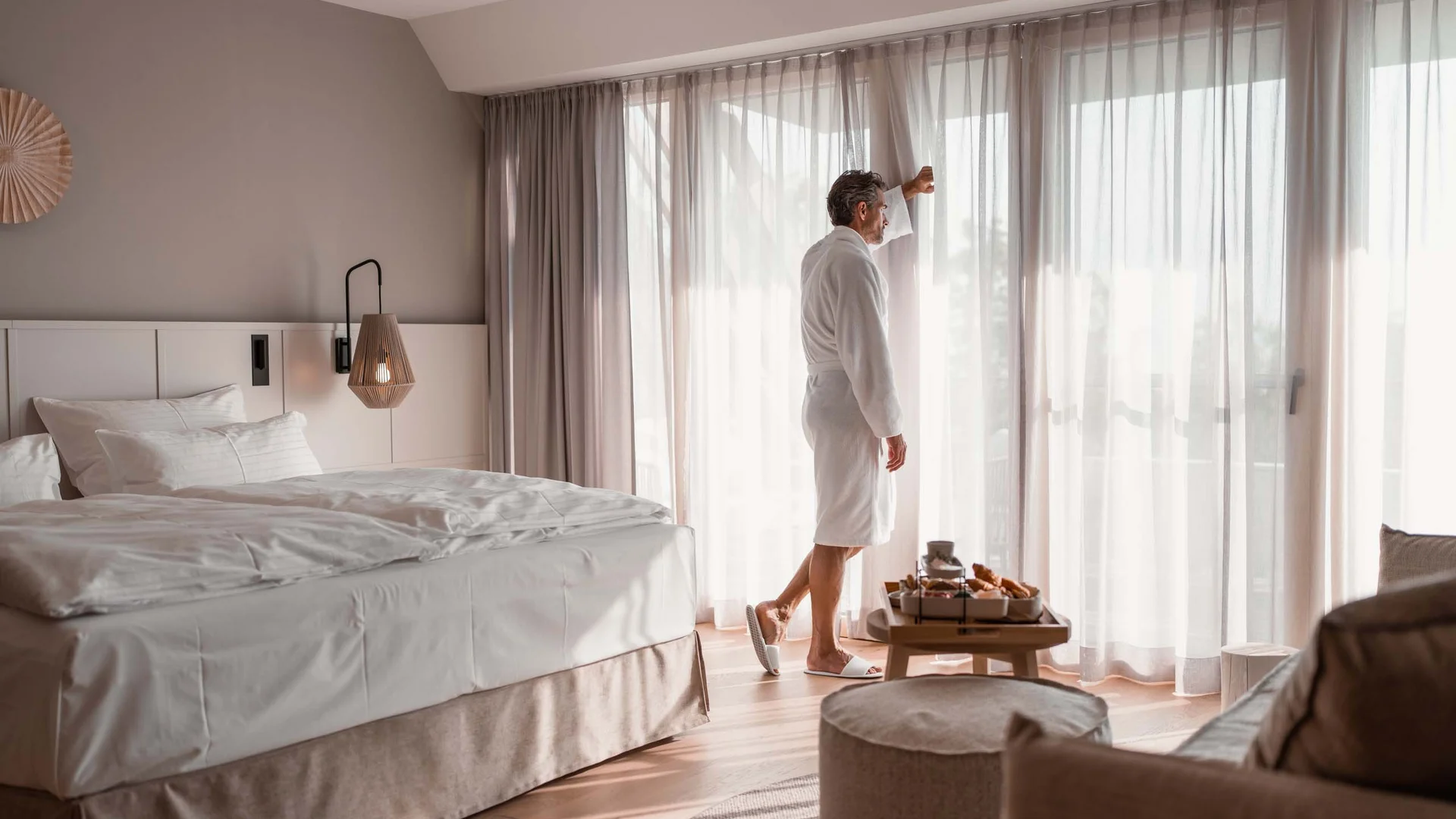 Mann im Bademantel schaut durch Vorhänge in einem hellen Hotelzimmer